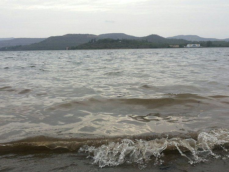 A wide shot of the Khadakwasla Dam, showing the water level rising high against the dam wall. The sky is cloudy, and the water appears choppy with small waves.